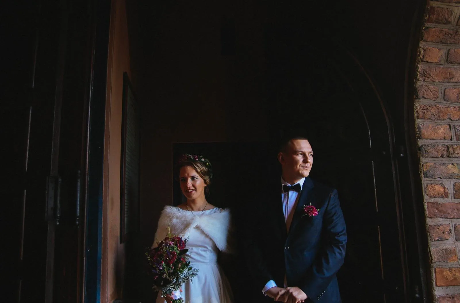 Para młoda stoi w mroku przy drzwiach kościoła, oświetleni naturalnym światłem – klasyczny portret po ceremonii.