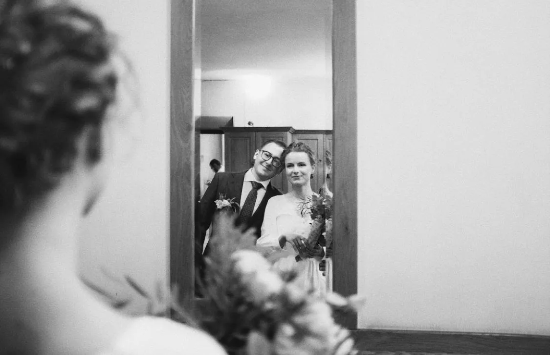 Para młoda uchwycona w lustrze – kobieta w sukni ślubnej z uśmiechem, mężczyzna z głową opartą o jej ramię.