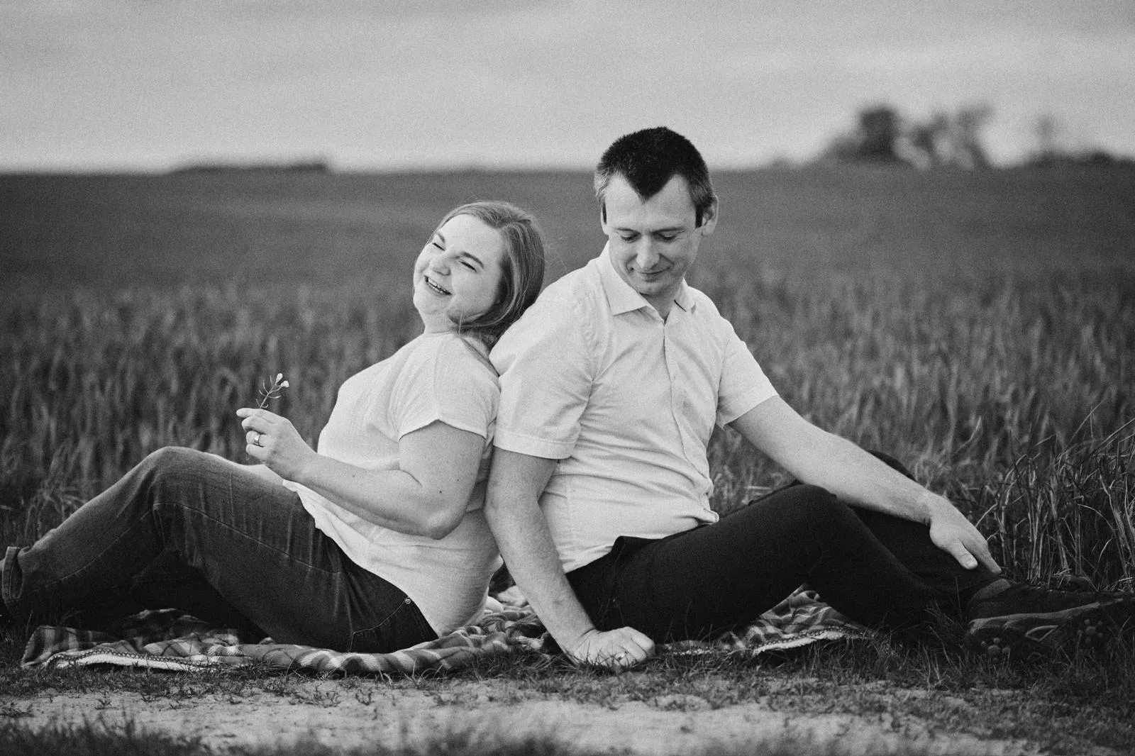 Para siedzi plecami do siebie na kocu w polu, sesja narzeczeńska na łonie natury.
