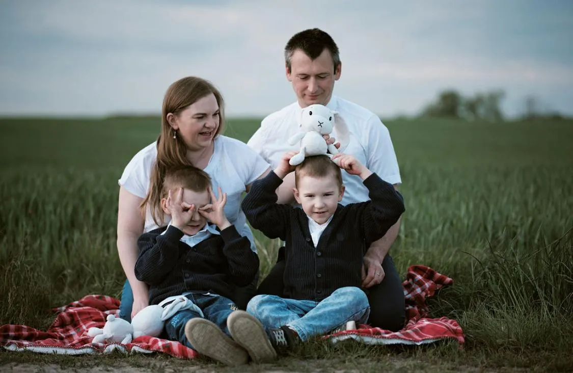 Rodzina siedzi na kocu w polu, dzieci robią zabawne miny i bawią się maskotką – spontaniczna i radosna scena.